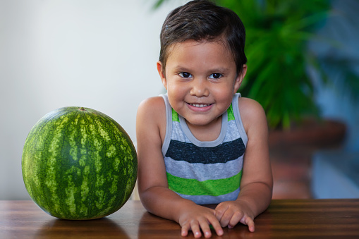 A cute young boy sitting next to a ripe watermelon he will be eating as part of a low-calorie and nutritious treat.