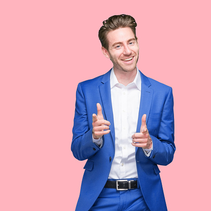 Waist up of aged 20-29 years old who is slim with short hair caucasian young male businessman standing in front of colored background wearing button down shirt who is smiling and showing finger gun who is pointing