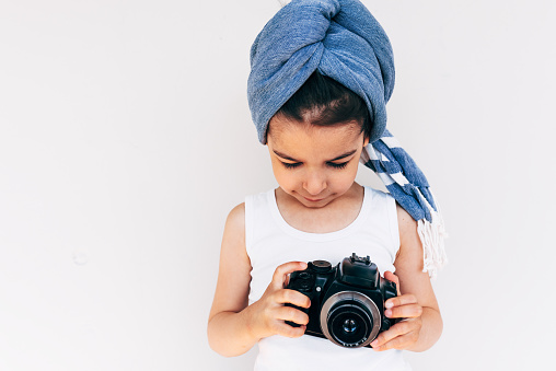 Image of cute little girl wearing turban on the head holding an photo camera, isolated on white background. Adorable child taking a picture with a professional camera posing on white background.