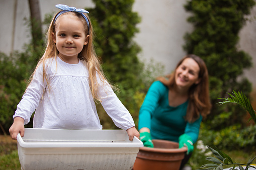 Shot of adorable toddler girl smiling and looking at camera while holding a flower pot and assisting her mother in planting flowers in their back yard.