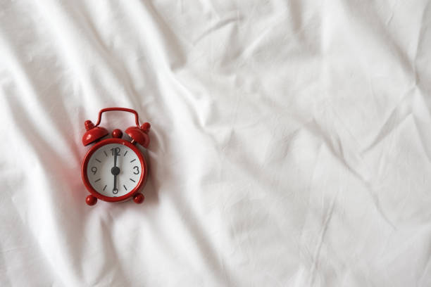 Red analog clock on white rumpled sheets. Top view, flat lay, copy space. Horizontal. Concept of awakening, sleep, early rise, productivity, productive day. For social media, blog. Minimal style