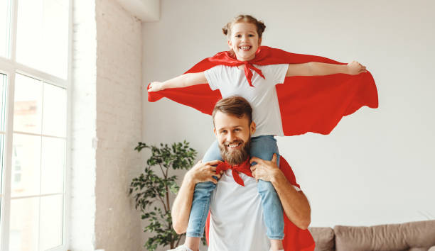 Positive father and daughter spending time together at home Smiling bearded young man holding cute girl on shoulders while playing superheroes together dressed in red superhero cloak in light room cape garment stock pictures, royalty-free photos & images