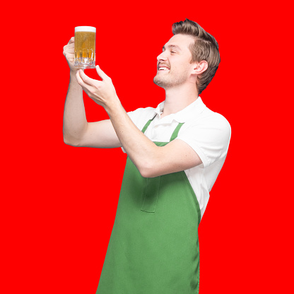 Inspector / inspect / -  portrait of with brown hair caucasian male standing in front of colored background wearing apron who is looking who is examining and holding drink