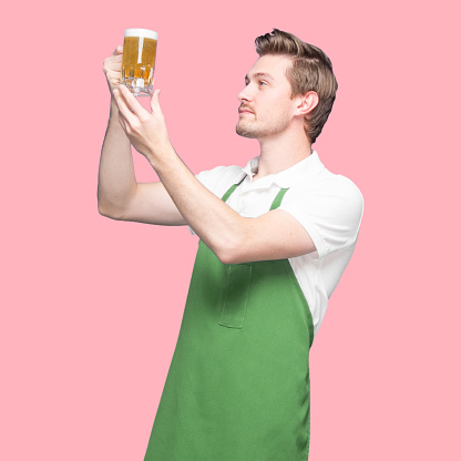 Inspector / inspect / -  one person of with short hair caucasian young male standing in front of colored background wearing polo shirt who is looking who is examining and holding craft beer