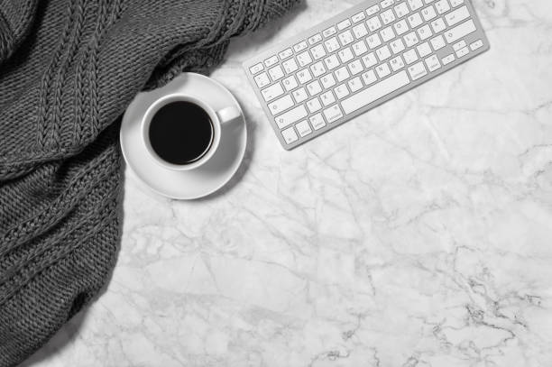 istock Cozy workspace with marble desk, warm wool plaid or scarf, coffee cup and keyboard in black and white 1224468018