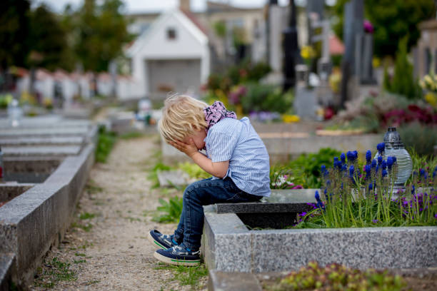 little toddler boy, sitting on a grave in cemetery, sad and lonely, springtime - cemetery child mourner death imagens e fotografias de stock