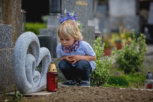 Little toddler boy, sitting on a grave in cemetery, sad and lonely
