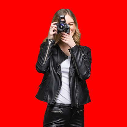 Full length of aged 20-29 years old who is beautiful with blond hair generation z young women photographer standing in front of colored background wearing pants who is laughing who is photographing and holding camera