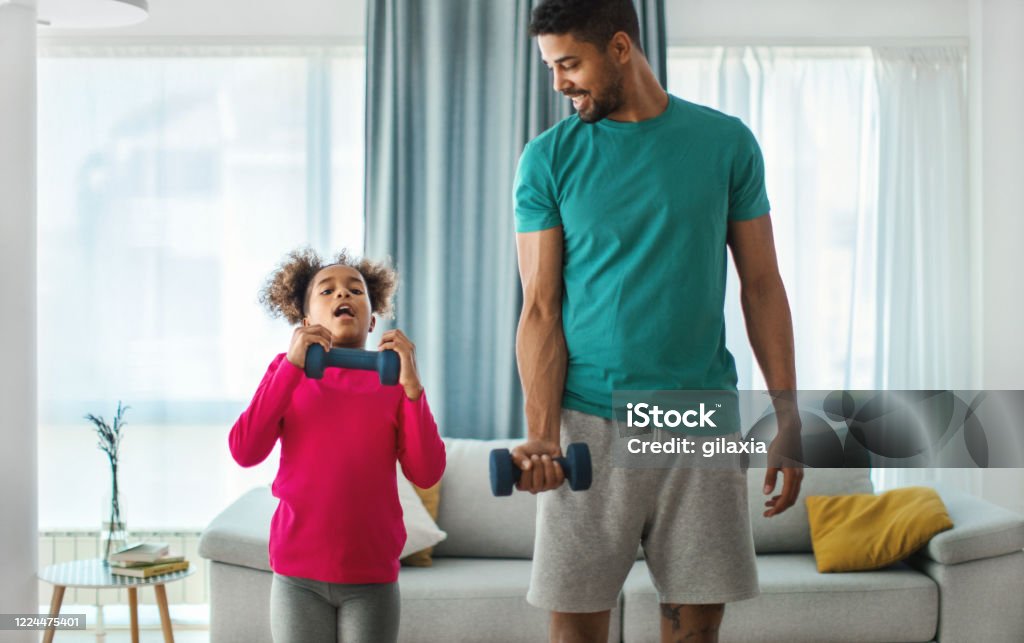 Father and daughter exercising at home. Closeup front view of a young african american man exercising at home. They are lifting some lightweight dumbbells. The girls is struggling with the exercise and desperately looking at th camera. Father Stock Photo