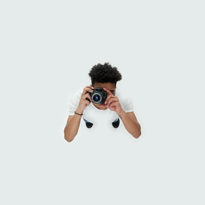 Directly above view of aged 18-19 years old with curly hair african-american ethnicity young male photographer standing in front of white background wearing shirt who is smiling who is photographing and holding camera