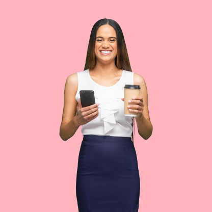 Front view of aged 30-39 years old who is beautiful african ethnicity female business person standing in front of colored background wearing business casual who is cheerful and holding coffee cup and using smart phone