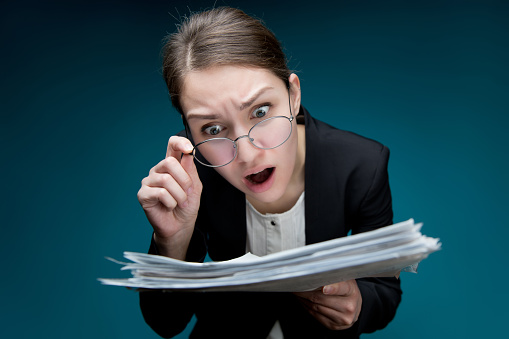 A young woman puts glasses on her nose and looks at documents with angry surprise. Director or teacher dissatisfied with paperwork or errors in them