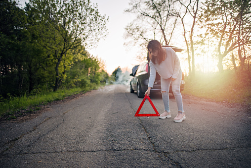 Young and beautiful woman placing the warning triangle symbol in front of her broken down car. She looks worried and sad because she is away from city and it is getting dark