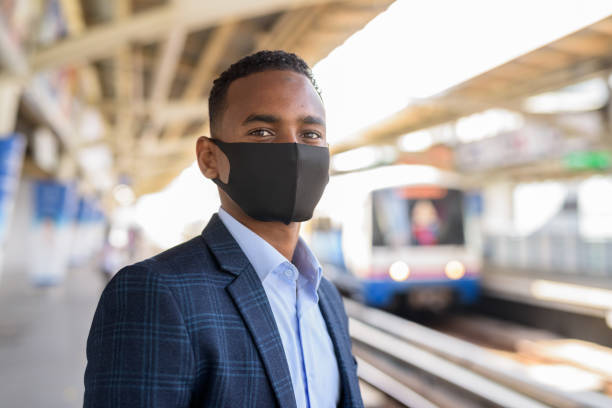 Young African businessman with mask for protection from corona virus outbreak thinking and waiting at sky train station Portrait of young African businessman with mask for protection from corona virus outbreak at the sky train station bts skytrain stock pictures, royalty-free photos & images