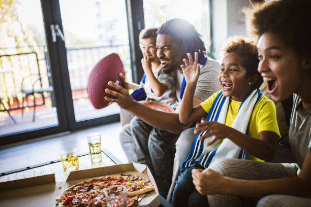 Cheerful black family cheering while watching rugby match on TV at home. Young joyful black family having fun while cheering for their favorite American football team at home. american football sport stock pictures, royalty-free photos & images