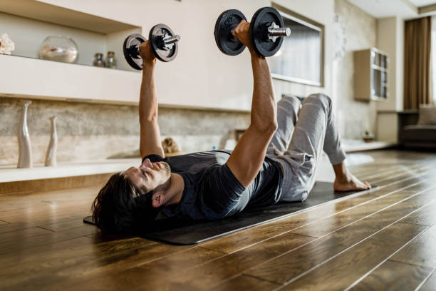 Muscular build man exercising strength with weights on a floor. Determined athletic man exercising with hand weights on a floor at home. Copy space. weightlifting stock pictures, royalty-free photos & images