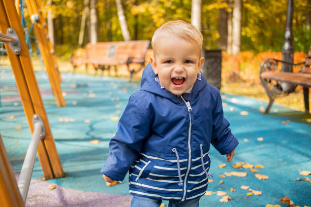 verticale d’un petit garçon blond mignon d’enfant en bas âge dans une veste bleue regardant l’appareil-photo et riant heureusement. en arrière-plan une aire de jeux dans le parc par une journée ensoleillée dans le flou. plan rapproché, mise au  - playground cute baby blue photos et images de collection