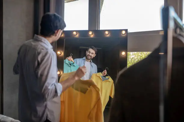 Photo of Man reflecting in the mirror with tshirts in his hands