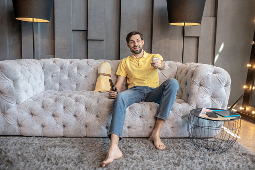 Free time. Young adult man barefoot in jeans and a yellow tshirt sitting on a sofa with a tv remote, smiling