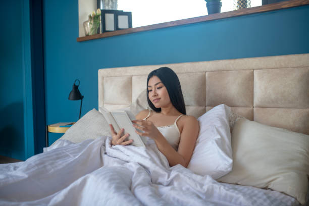 istock Young brunette sitting in bed in night dress, reading book 1224537567