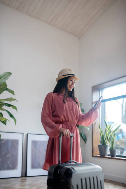istock Dark-haired female in straw hat standing, holding suitcase handle and checking her mobile phone 1224537630