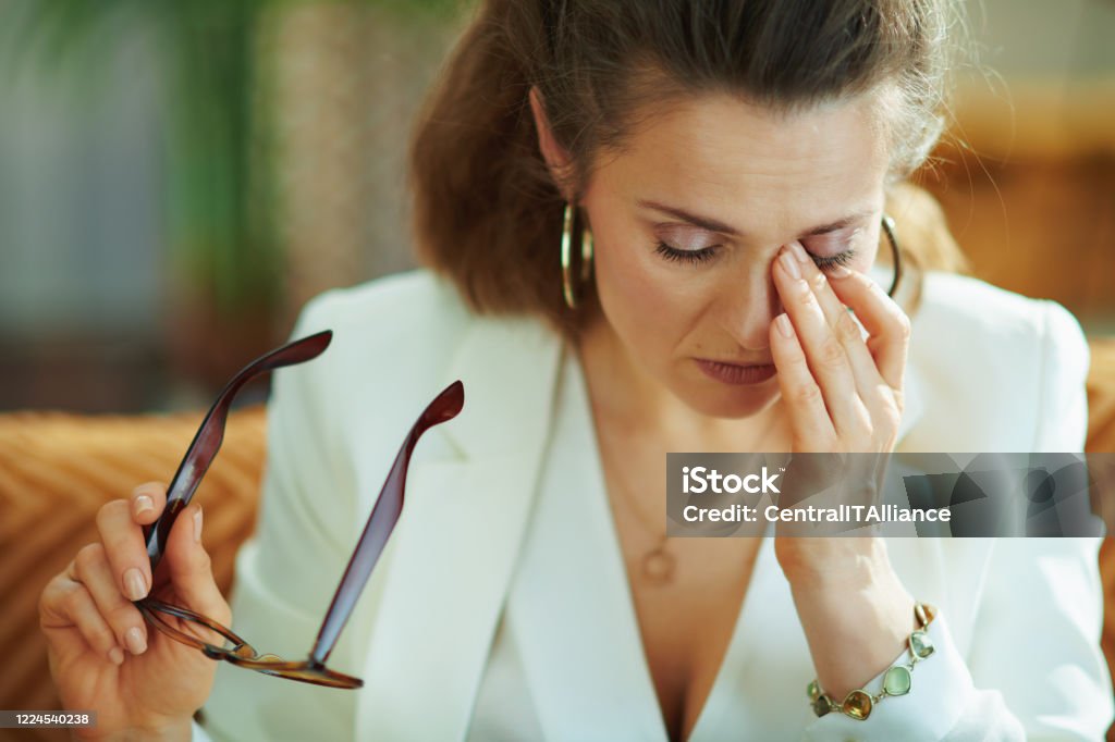 tired elegant housewife glasses rubbing eyes tired elegant middle age housewife in white blouse and jacket with glasses rubbing eyes in the modern living room in sunny day. Eye Stock Photo