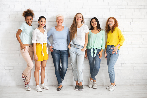 Diverse Ladies Of Different Age Posing Standing Over White Brick Wall Smiling To Camera. Full Length