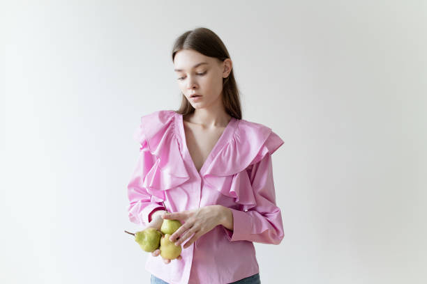 Beautiful young woman with nude color make-up wearing pink shirt and blue jeans holding 3 pears on white background Portrait of beautiful young woman with nude color make-up wearing pink shirt and blue jeans holding three pears on white background Perfect glowing skin Fashion model studio portrait perfect pear stock pictures, royalty-free photos & images