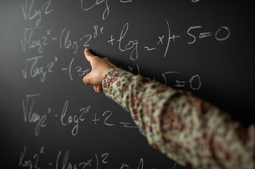 Hand of female math teacher showing solution for math problem on blackboard, close-up