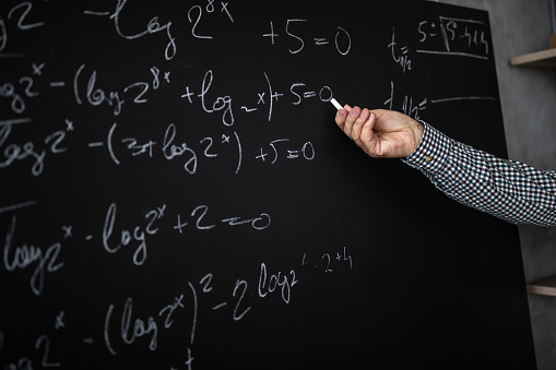 Math teacher standing beside blackboard, holding chalk and explaining math formula to his online students