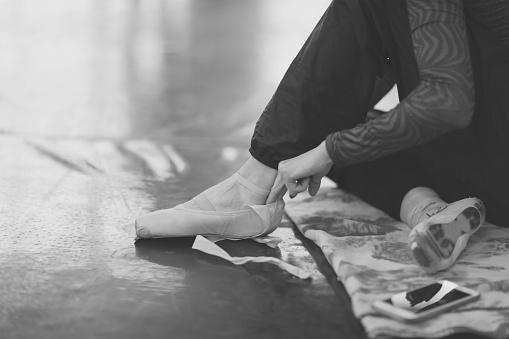 Ballerina sits on the floor in the theater's rehearsal hall and puts on pointe shoes