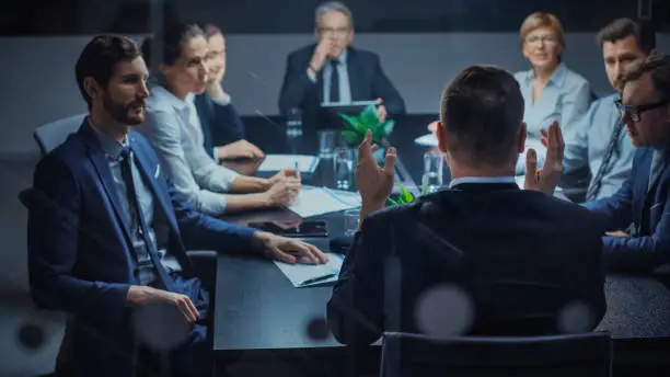 Photo of Late at Night In the Corporate Office Meeting Room: At Conference Table Executive Director Talks to a Board of Directors, Investors and Business Associates. Over the Shoulder Shot.