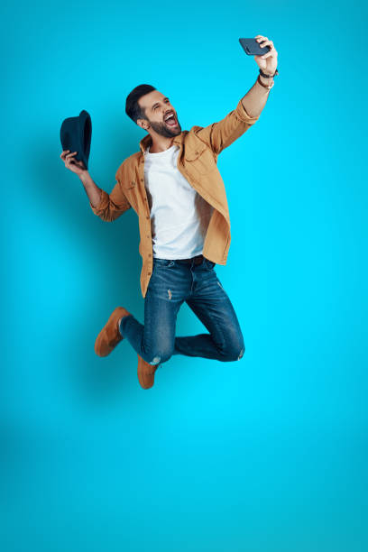 istock Full length of handsome young man smiling and taking selfie while hovering against blue background 1224564249