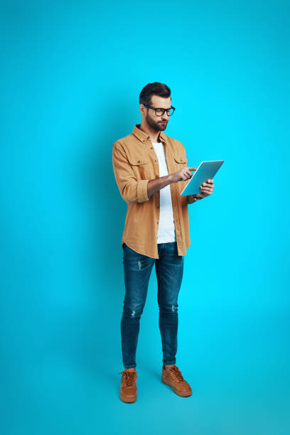 istock Full length of young man in smart casual wear using digital tablet while standing against blue background 1224564323