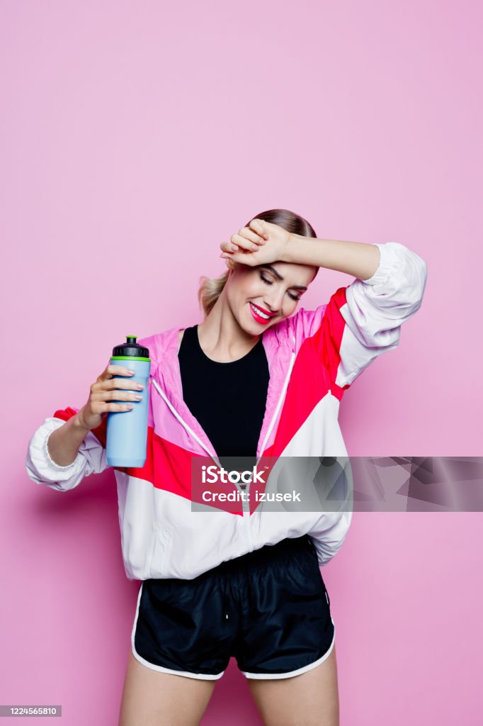 80's style portrait of smiling woman in sports clothes against pink background Sport portrait of beautiful woman wearing oversized tracksuit and black shorts. Happy woman holding water bottle. Studio shot, pink background. Exercising Stock Photo