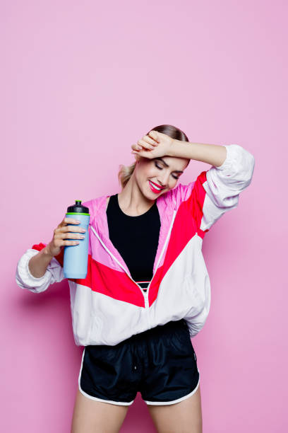 80's style portrait of smiling woman in sports clothes against pink background Sport portrait of beautiful woman wearing oversized tracksuit and black shorts. Happy woman holding water bottle. Studio shot, pink background. 80s aerobics stock pictures, royalty-free photos & images