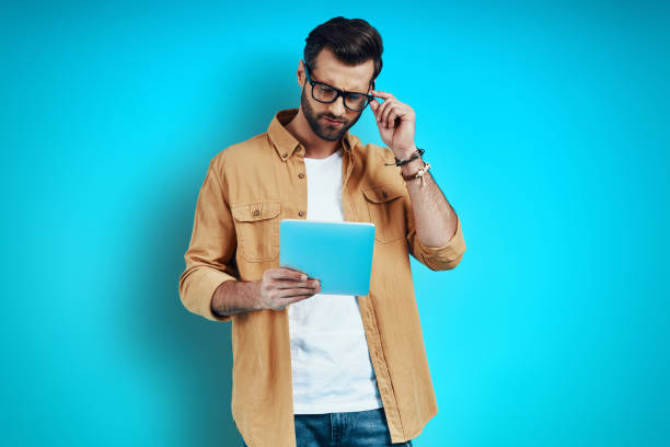 istock Thoughtful young man in smart casual wear using digital tablet and adjusting eyewear while standing against blue background 1224566062