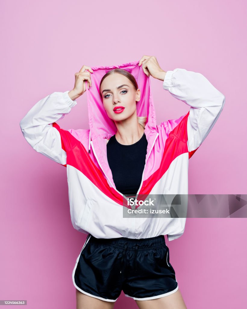 Fashion portrait of beautiful woman in hoodie tracksuit Mid adult beautiful woman wearing oversized 80’s style tracksuit and black shorts standing against pink background, smiling at camera. Retro Style Stock Photo