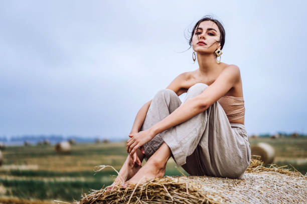 Brunette in linen pants and bare shoulders sitting on a hay bales in warm autumn day. Woman looking at camera. Behind her is a wheat field stock photo