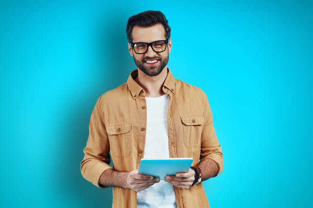 istock Young man in smart casual wear using digital tablet and looking at camera while standing against blue background 1224568352