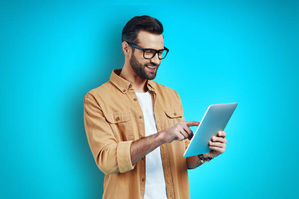 istock Busy young man in smart casual wear using digital tablet while standing against blue background 1224568506