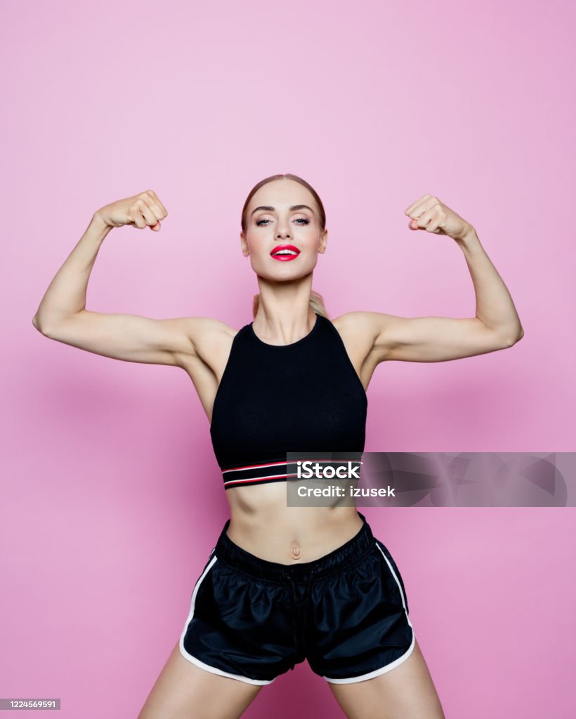 Portrait of slim woman in sports clothing against pink background Sport portrait of beautiful woman wearing sport black shorts and top, flexing her muscles. Studio shot, pink background. 30-34 Years Stock Photo