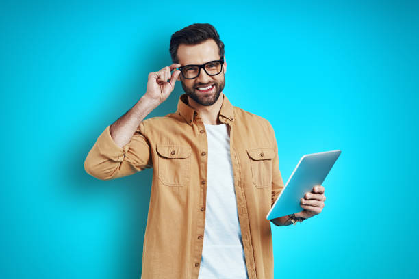 istock Confident young man in smart casual wear using digital tablet and adjusting eyewear while standing against blue background 1224569759