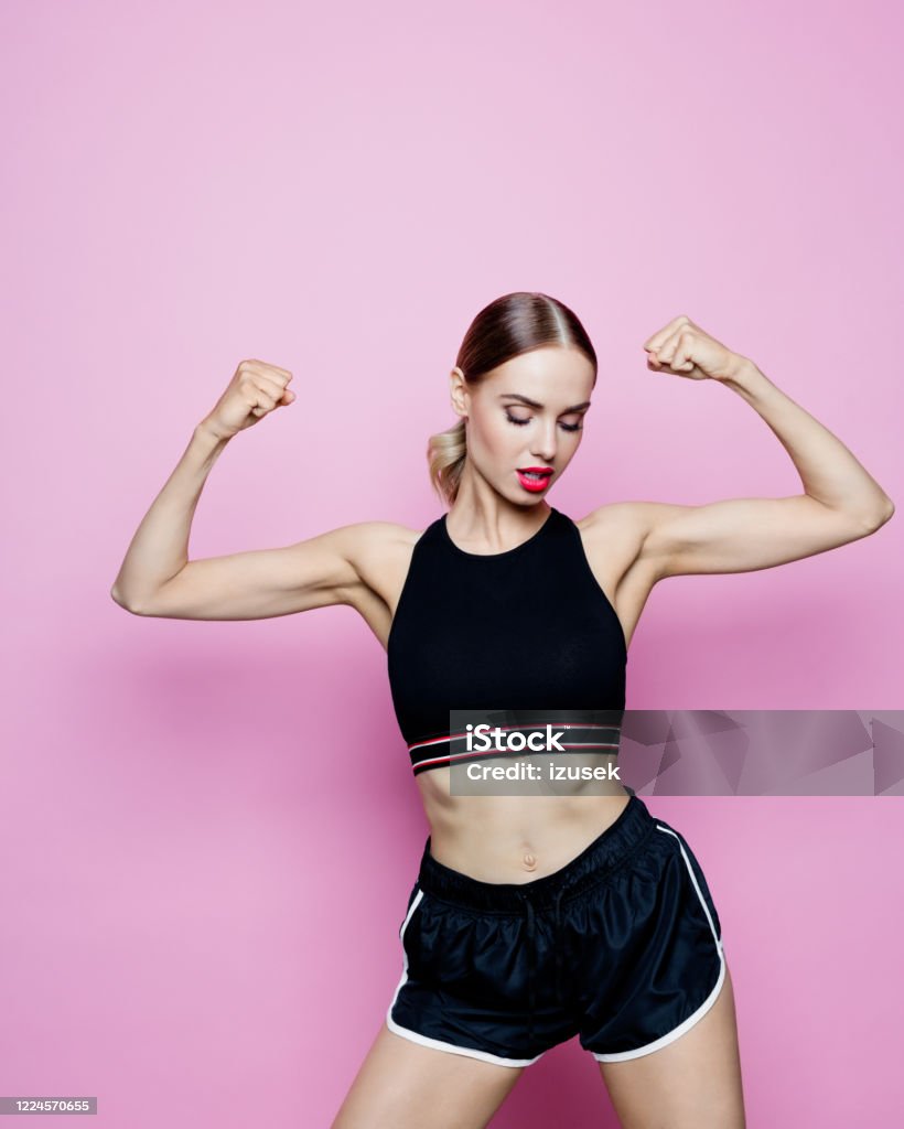 Portrait of strong woman in sports clothing against pink background Sport portrait of beautiful woman wearing sport black shorts and top, flexing her muscles. Studio shot, pink background. 30-34 Years Stock Photo