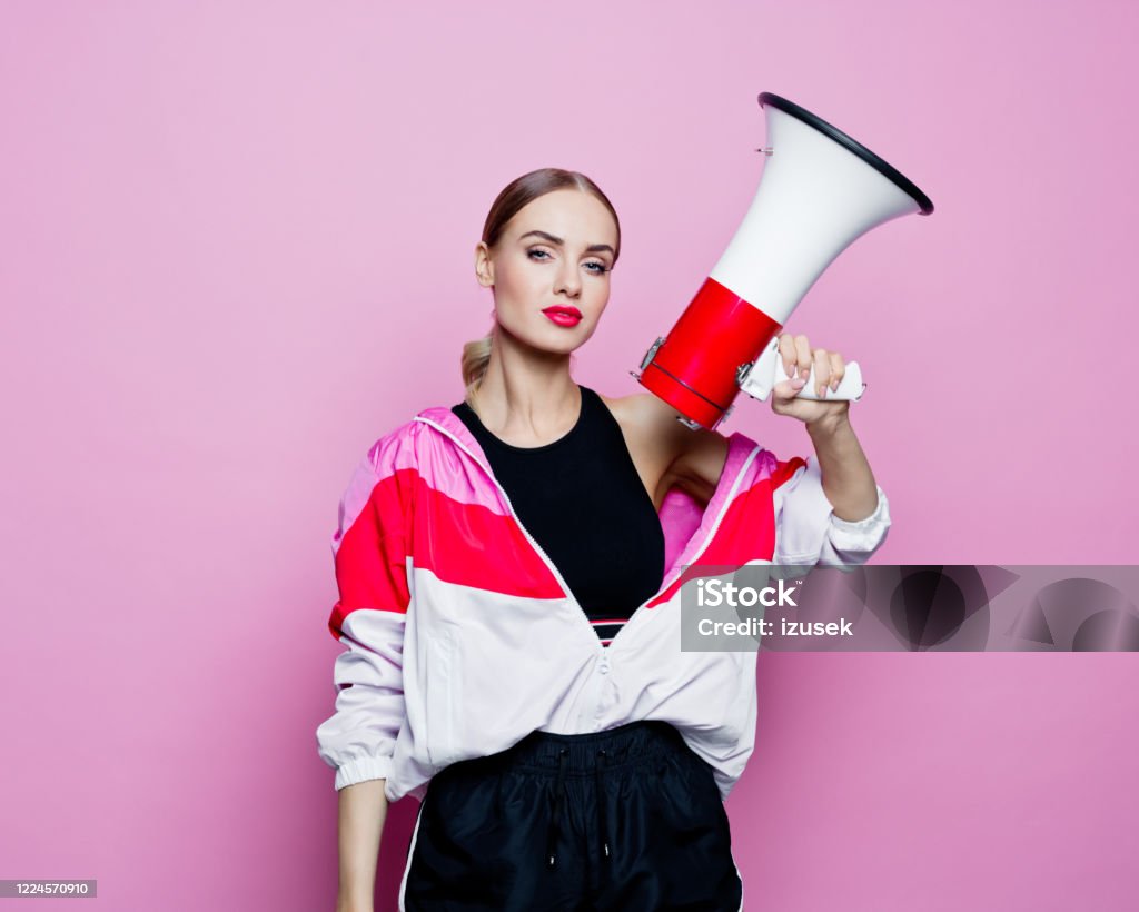 Sports portrait of beautiful woman in 80's style tracksuit holding megaphone Mid adult beautiful woman wearing oversized tracksuit and black shorts and top standing against pink background, looking at camera. Confident woman holding megaphone. Megaphone Stock Photo