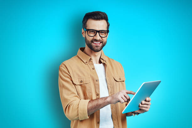 istock Busy young man in smart casual wear using digital tablet and looking at camera while standing against blue background 1224570933