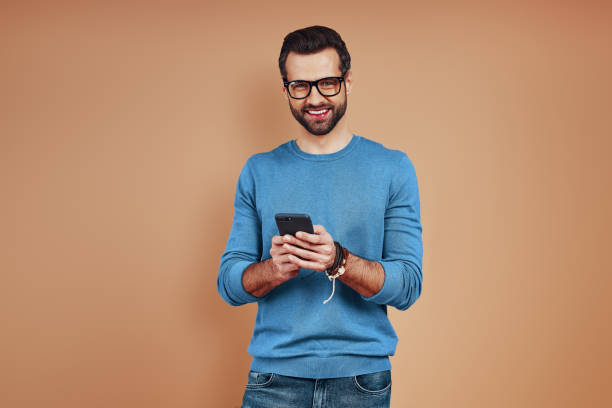 istock Happy young man in casual wear using smart phone and smiling while standing against brown background 1224571856
