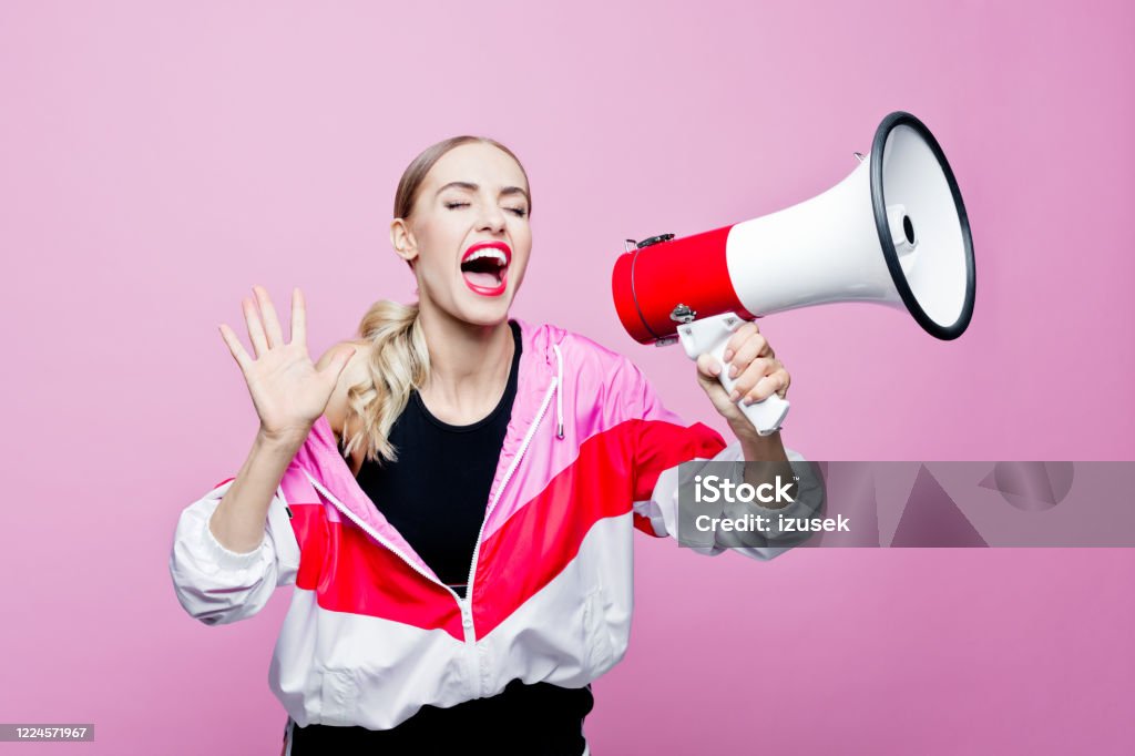 Sports portrait of woman in tracksuit shouting into megaphone Mid adult beautiful woman wearing oversized tracksuit and black shorts and top standing against pink background. Confident woman shouting into megaphone. Public Speaker Stock Photo