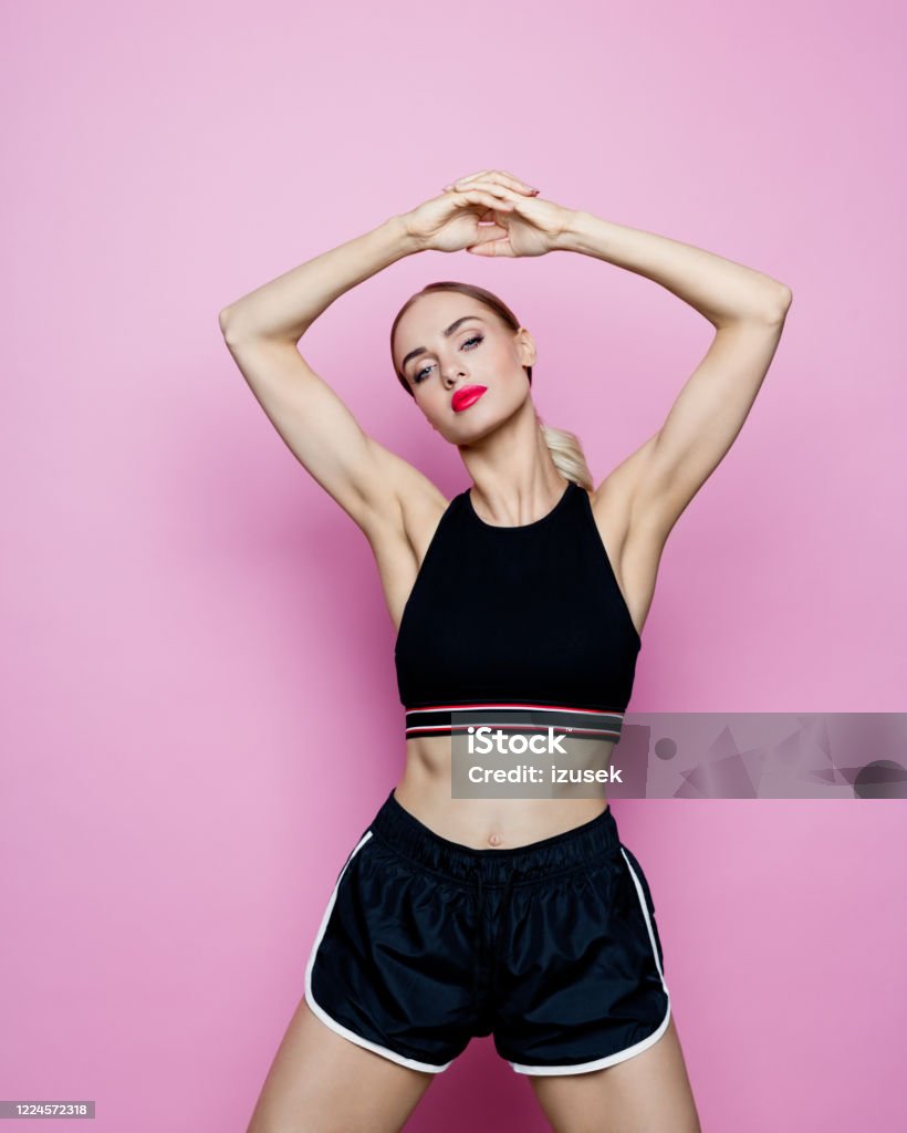 Studio portrait of slim woman in sports clothing against pink background Sport portrait of beautiful woman wearing sport black shorts and top, standing with raised hands. Studio shot, pink background. Women Stock Photo