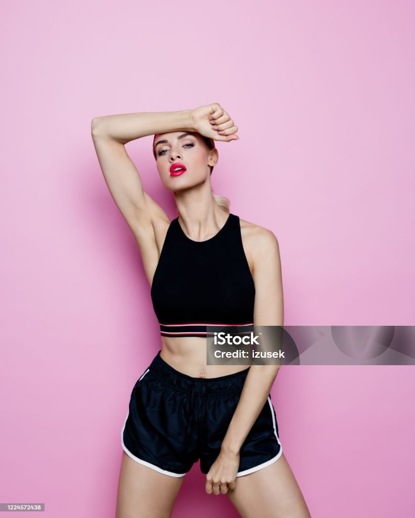Studio portrait of slim woman in sports clothing against pink background Sport portrait of beautiful woman wearing sport black shorts and top, standing with raised hand. Studio shot, pink background. Beautiful Woman Stock Photo